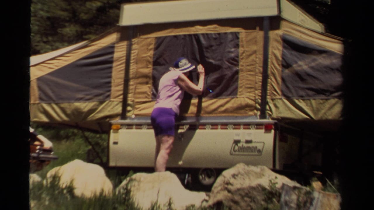 In 1980 Mendocino camping footage, a woman in a pink top and purple shorts works beside a tan pop-up camper among rocks and trees.