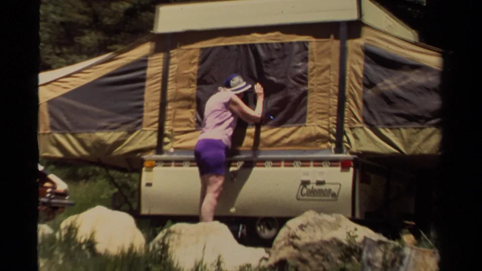 In 1980 Mendocino camping footage, a woman in a pink top and purple shorts works beside a tan pop-up camper among rocks and trees.