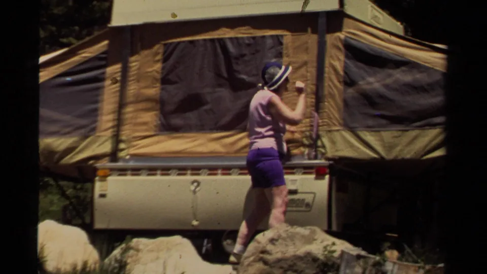 The woman continues adjusting the camper fabric or support near a tan Coleman pop-up camper in a wooded Mendocino campsite.