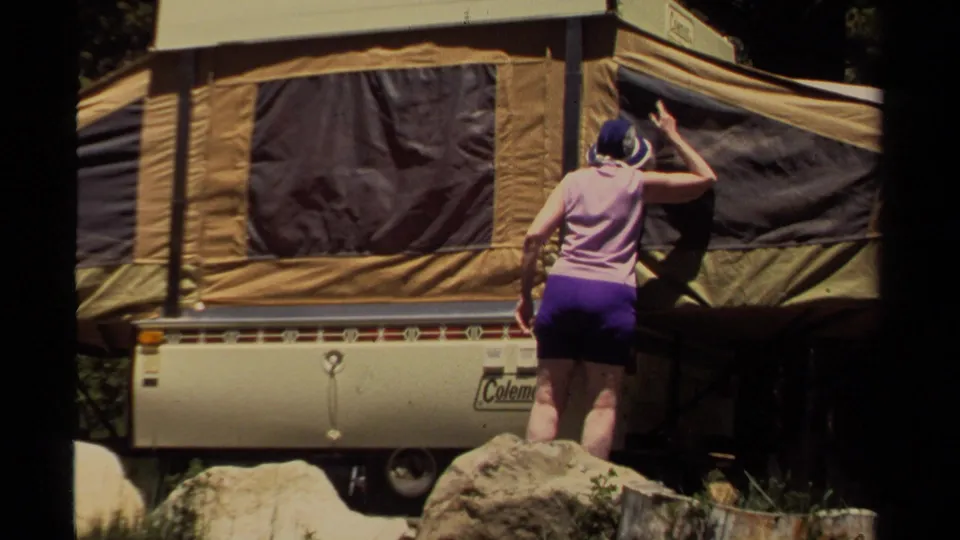 A rear view shows the woman reaching up to the camper side panel, with the tan trailer body, rocks, and tree shade visible.