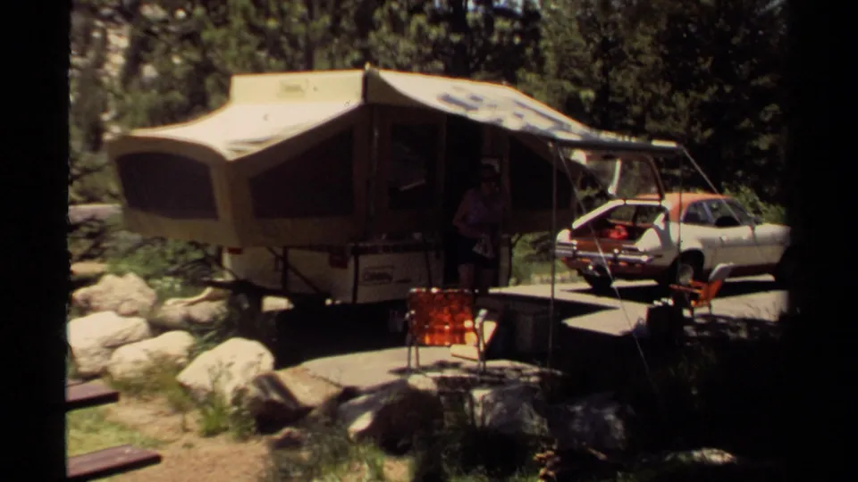 A wider 1980 Mendocino campsite view shows the opened pop-up camper, nearby car, folding chairs, rocks, and surrounding trees.