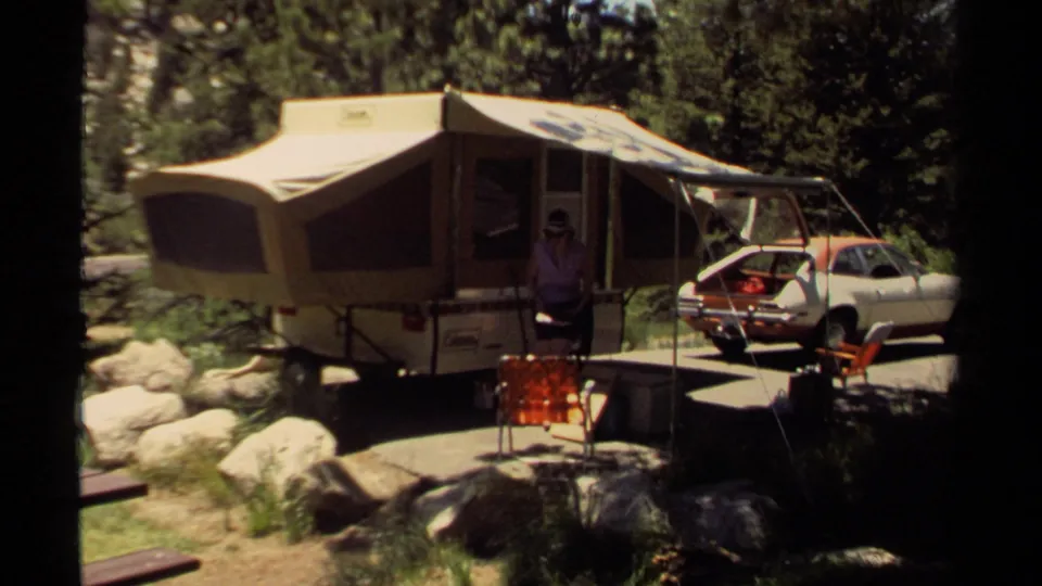 In Mendocino, California in 1980, a woman stands near a canvas pop-up camper, red folding chair, rocks, trees, and a parked compact car.