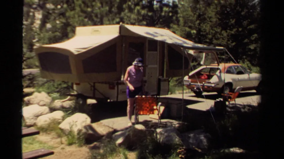 A woman in a purple shirt leans near a red folding chair at a 1980 Mendocino campsite with a pop-up camper and parked car behind her.