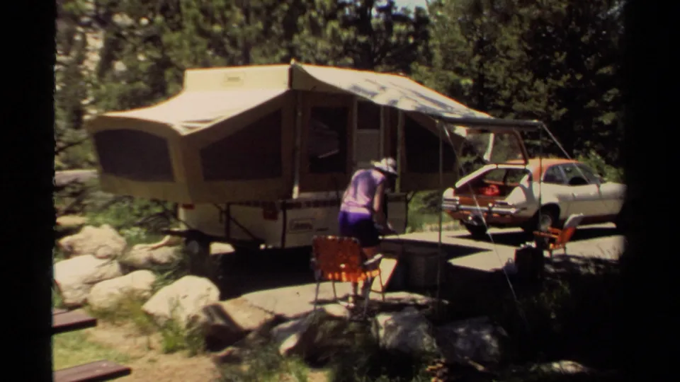 At the wooded Mendocino campsite, the woman bends near the red folding chair while the pop-up camper and compact car remain parked behind her.