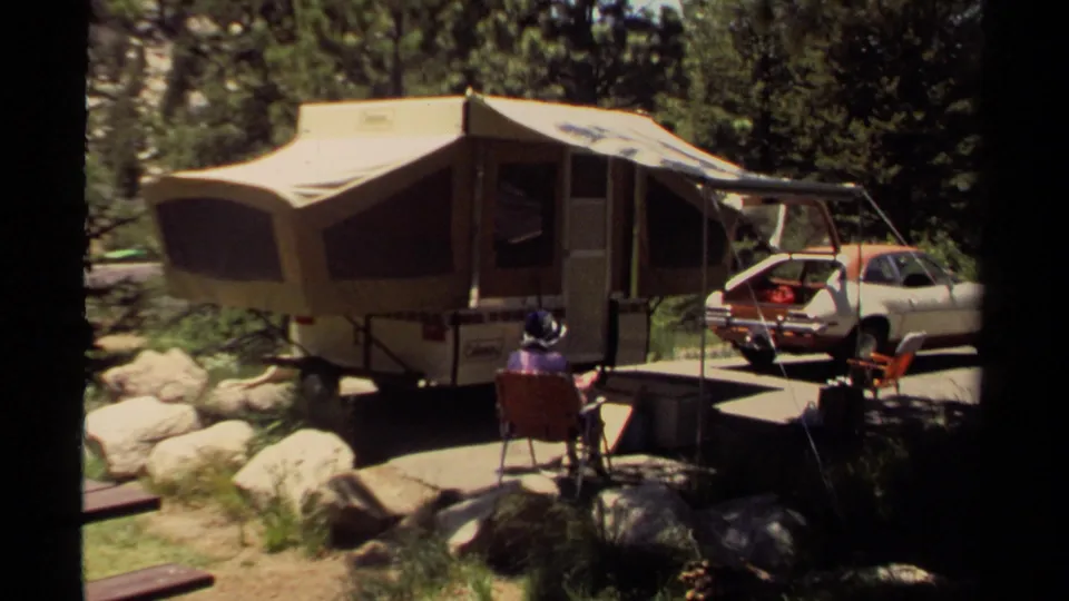 The woman sits in a red folding chair at the 1980 Mendocino campsite, facing the pop-up camper, awning, car, rocks, and trees.