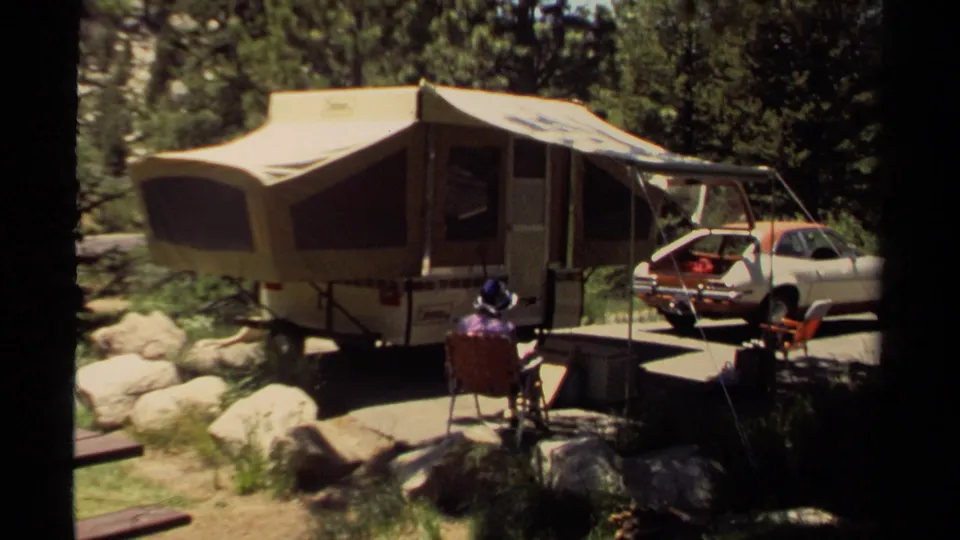 1980 Mendocino camping scene with a canvas pop-up camper, seated adult, folding chairs, trees, rocks, and a white and orange car.