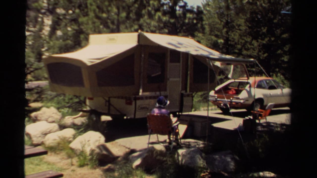Camper and parked car at a wooded campsite with a person seated in front.