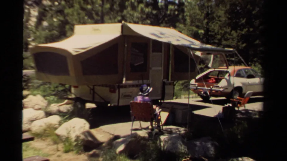 Wide 1980 Mendocino home-movie frame of a canvas pop-up camper, seated adult, car, rocks, chairs, and shaded trees.