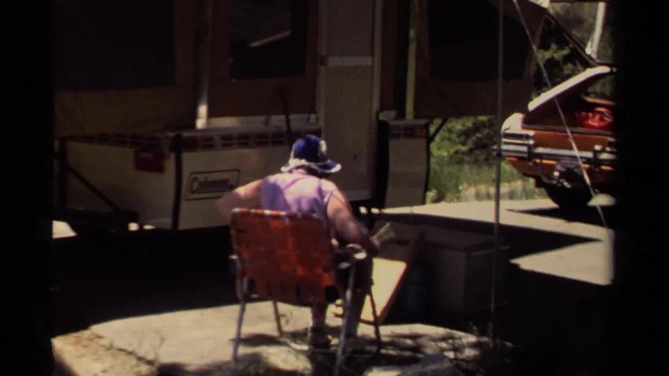 Closer 1980 Mendocino campsite frame with an adult in a blue hat seated near a pop-up camper, awning, car, and folding furniture.
