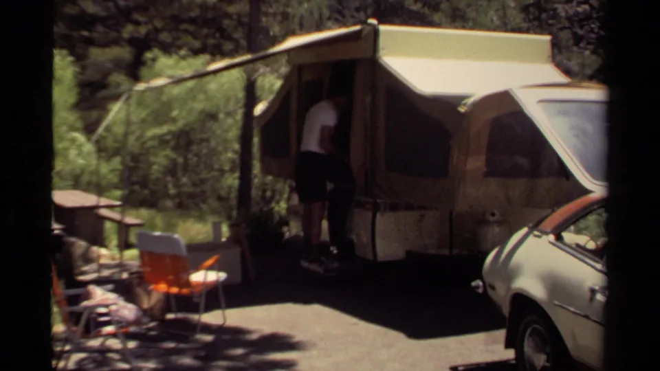 1980 Mendocino home-movie frame showing a man entering or adjusting a pop-up camper at a shaded wooded campsite.