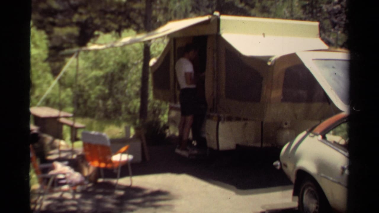 Wide 8mm frame of a man beside a pop-up camper in a Mendocino wooded campsite, with folding chairs and a car visible.