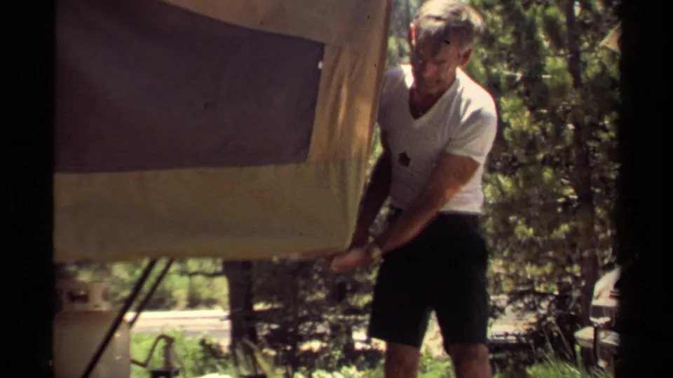 Closer 1980 home-movie view of a man in a white t-shirt and dark shorts working on the side of a pop-up camper.