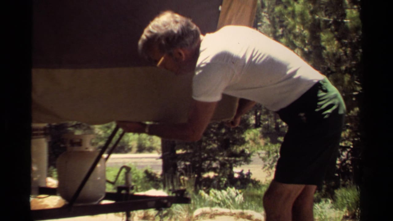 Man in a white T-shirt bends over a canvas-covered trailer at a wooded campsite.