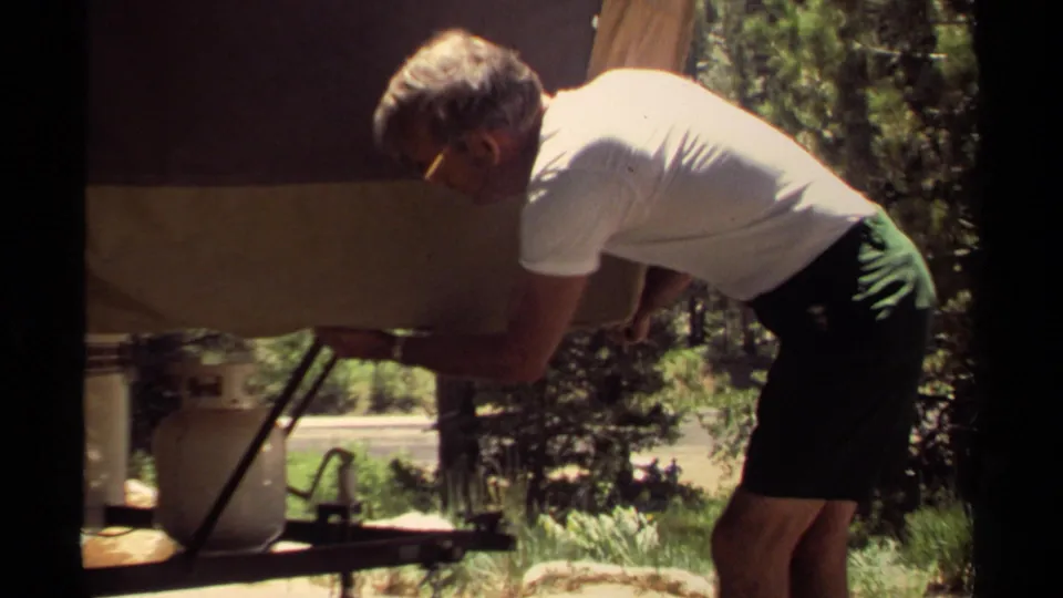 In 1980 Mendocino, California footage, a man bends at a small trailer while adjusting a canvas cover near trees and a propane tank.