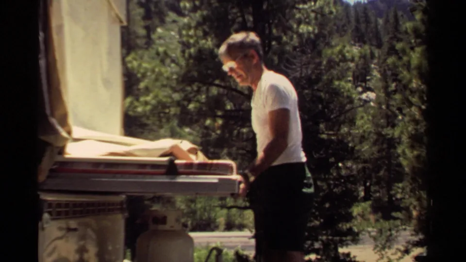 The 1980 Mendocino home-movie frame shows a man beside the trailer hardware, with canvas, trees, and a propane tank in view.