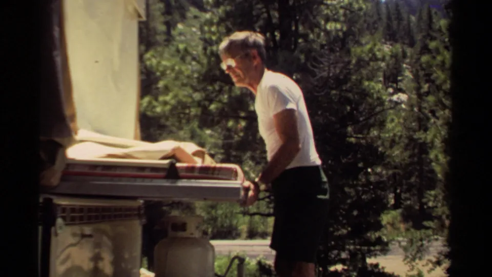 A man stands next to a small trailer in the wooded Mendocino campsite, framed by bright sunlight, trees, and visible 8mm film borders.