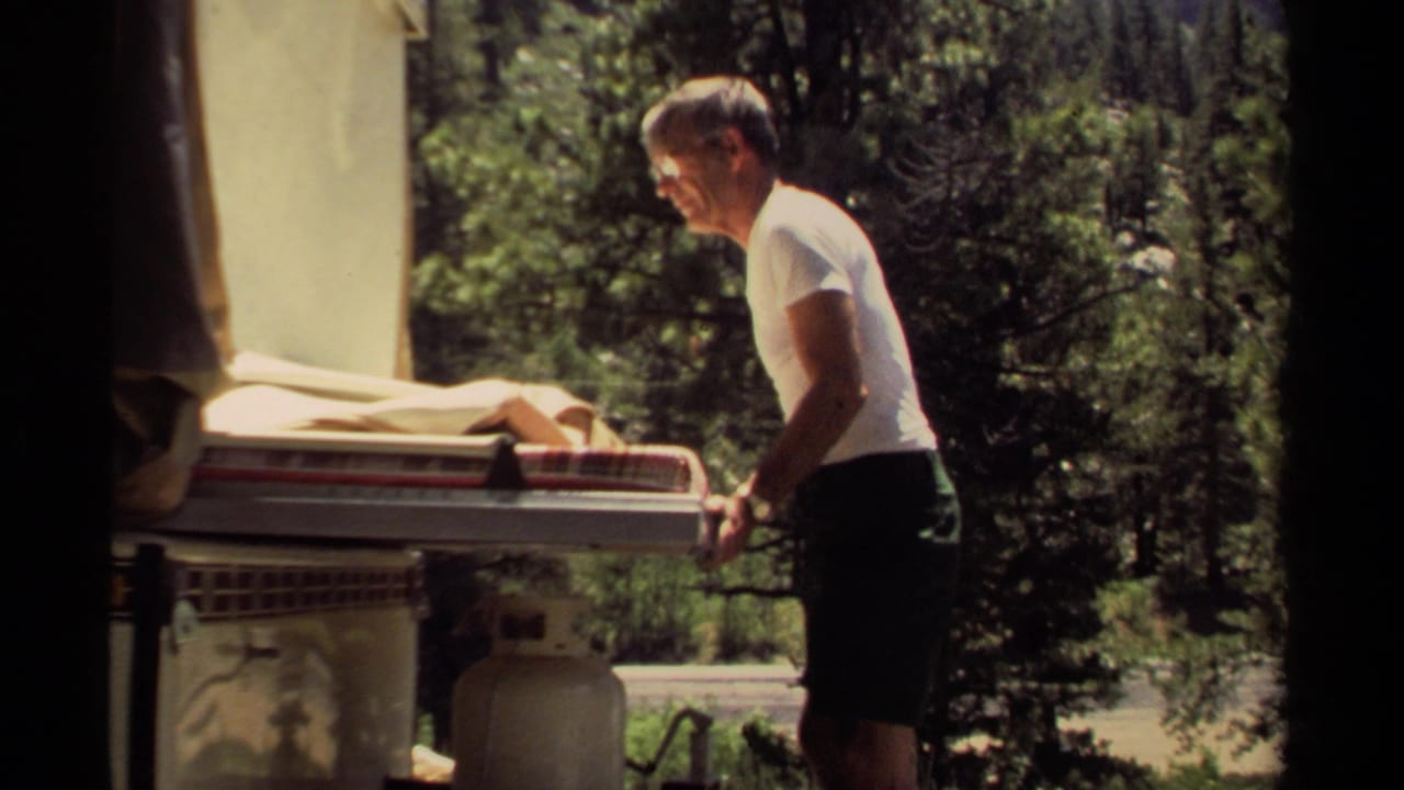 Man standing beside a camper trailer with mattresses and a propane tank in a wooded campground.