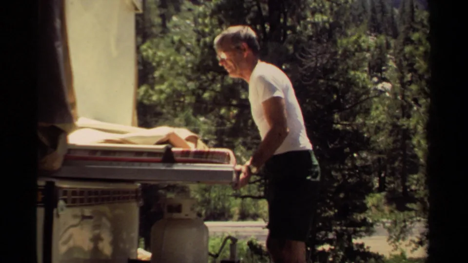 In Mendocino, California in 1980, a man stands beside a camper trailer with bedding pulled out under sunlit trees.