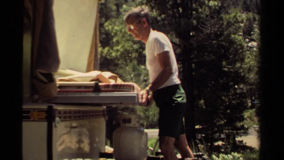 The man stands at the rear of the camper trailer in a wooded Mendocino campground, with bedding visible on the slide-out area.