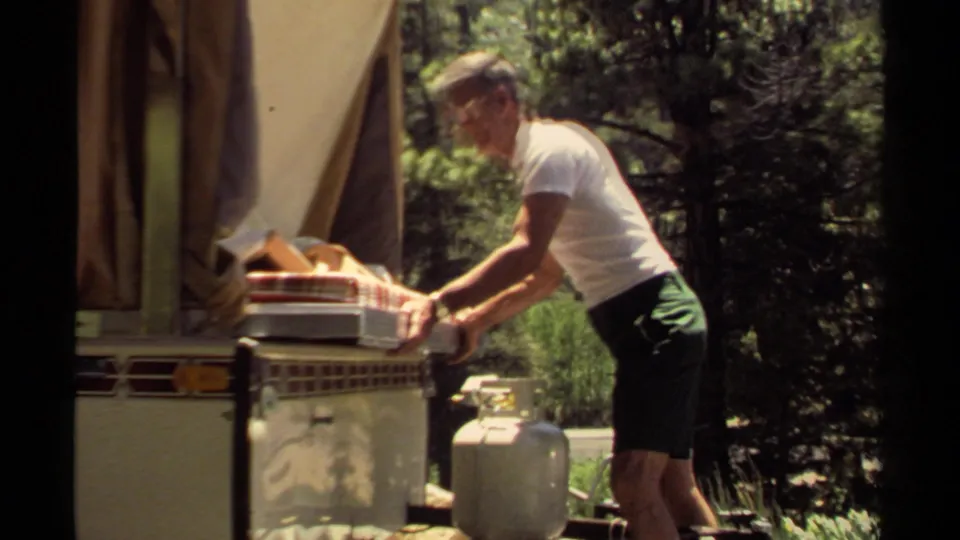 The man leans forward and pushes the mattresses into the camper trailer in the sunlit 1980 campground scene.