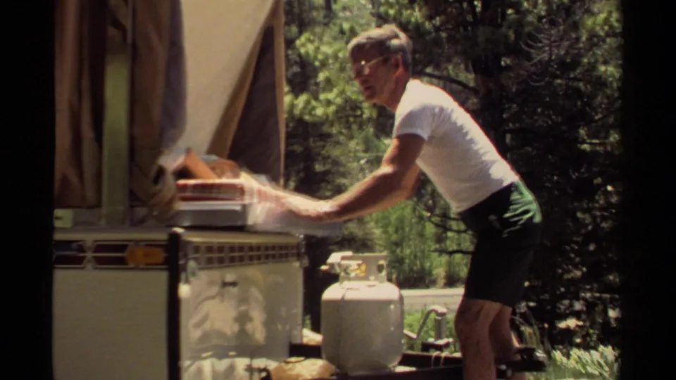 In 1980 Mendocino camping footage, a man leans toward a camper oven while handling a tray beside a propane tank and trees.
