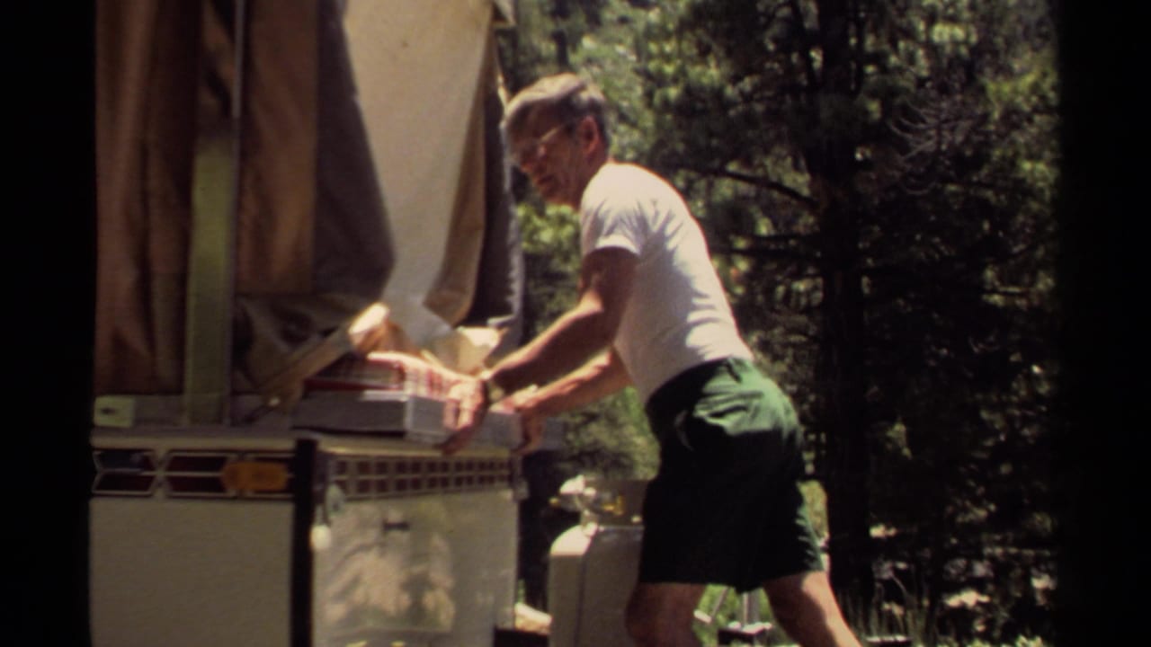 The man continues working the tray into the vintage camper oven, with the canvas awning and propane tank visible in the campsite.