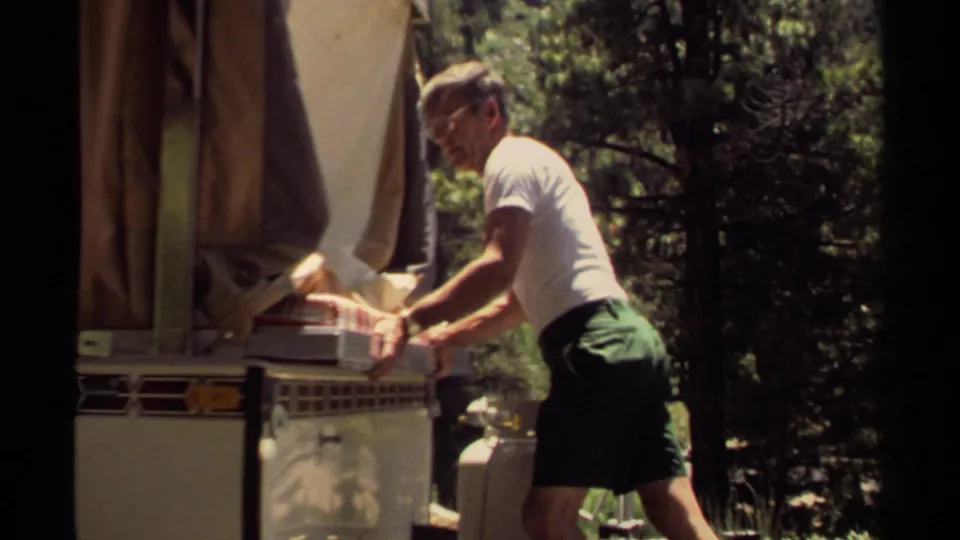 The man continues working the tray into the vintage camper oven, with the canvas awning and propane tank visible in the campsite.