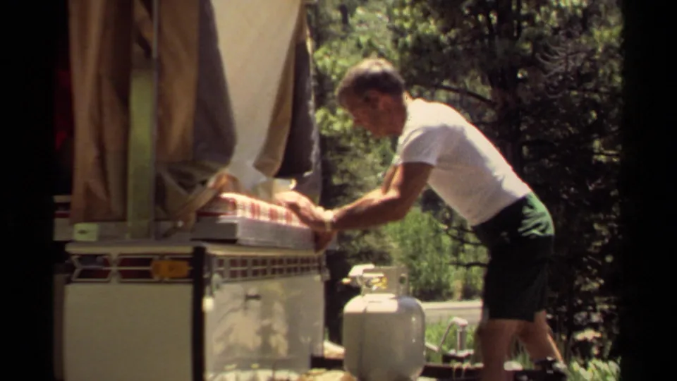 In the campsite setup, the man bends closer over the camper oven as he tries to slide the tray into place.