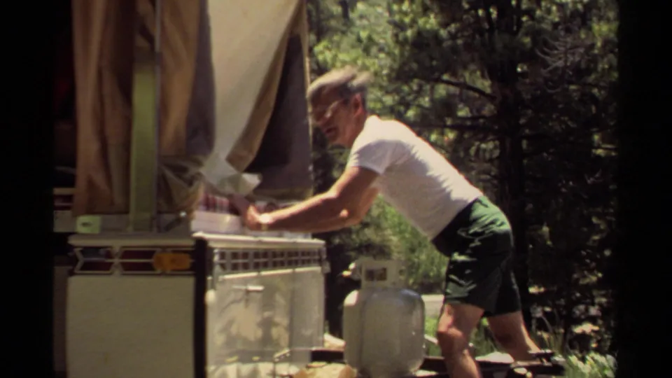 The man crouches and pushes the tray deeper into the camper oven, framed by canvas, trees, and campsite equipment.