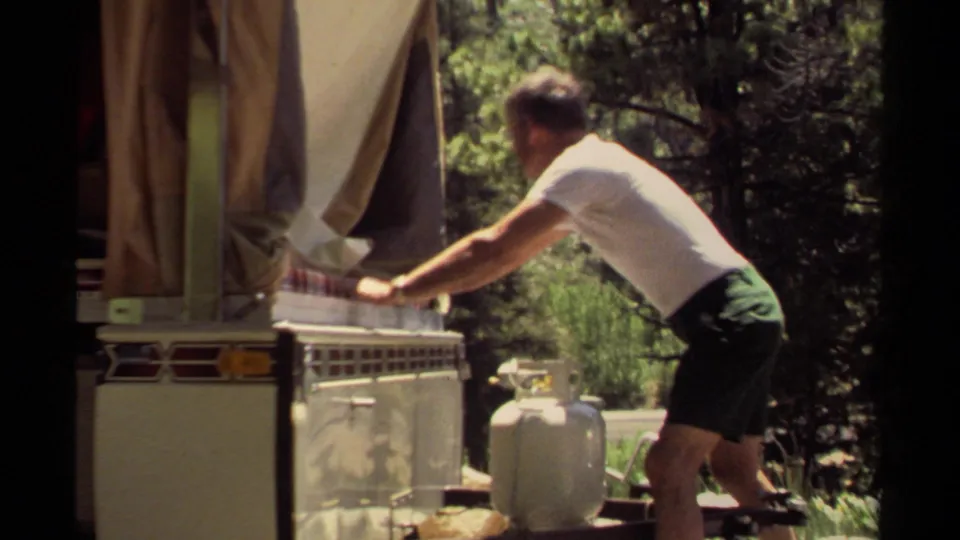 In Mendocino, California, 1980, a man in a white t-shirt works on a camper awning beside a propane tank in a sunny wooded campsite.
