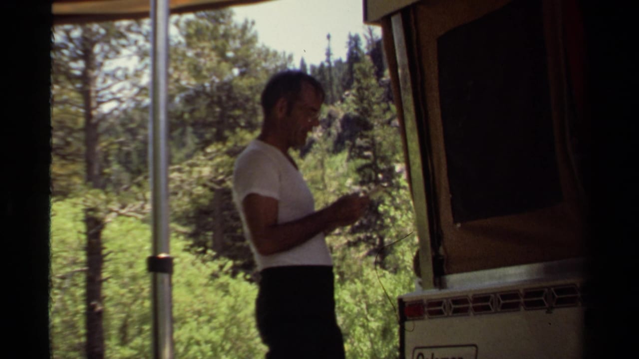 In the 1980 Mendocino camping scene, a man stands beside the camper trailer, looking down at something in his hands near a support pole and trees.