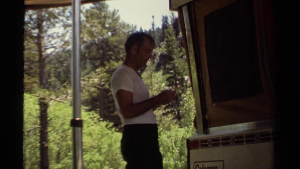 A man stands next to the Coleman camper in Mendocino, California, 1980, with trees and brush visible beyond the campsite.