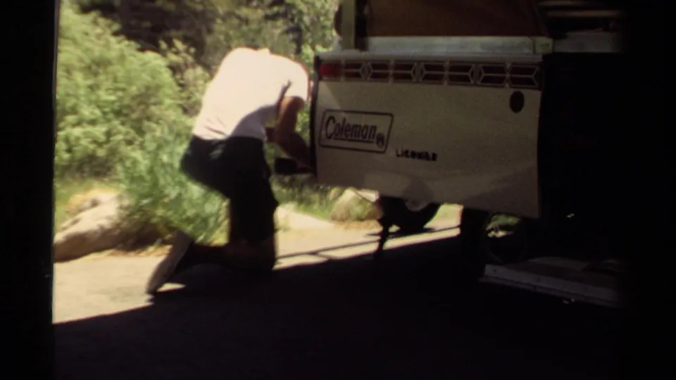 The man crouches near the rear or side of a Coleman camper trailer in the 1980 Mendocino campsite, with bright greenery beyond the shade.