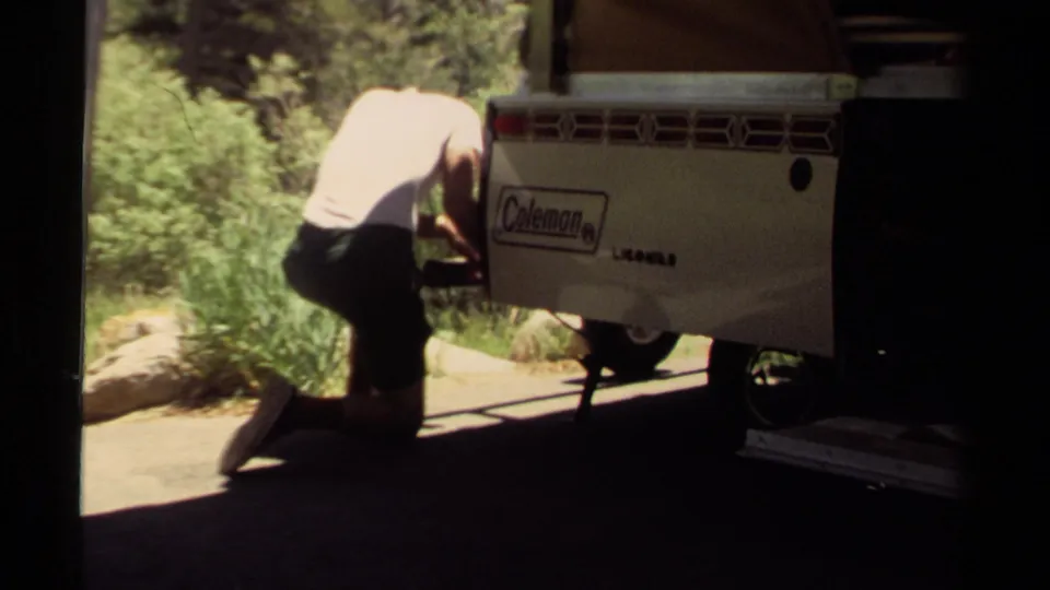 In 1980 Mendocino home-movie footage, a man kneels beside a Coleman camper trailer in a sunlit forest setting.