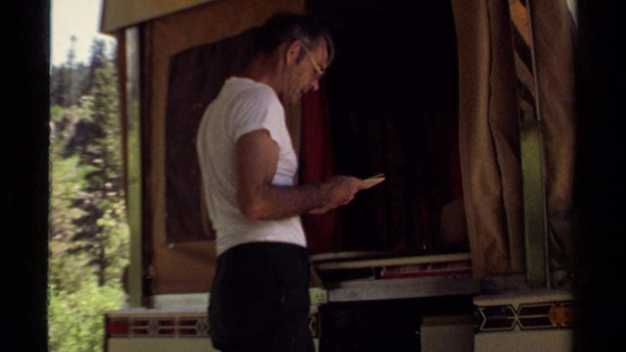 Man stands beside the open camper trailer while looking down at a small booklet.