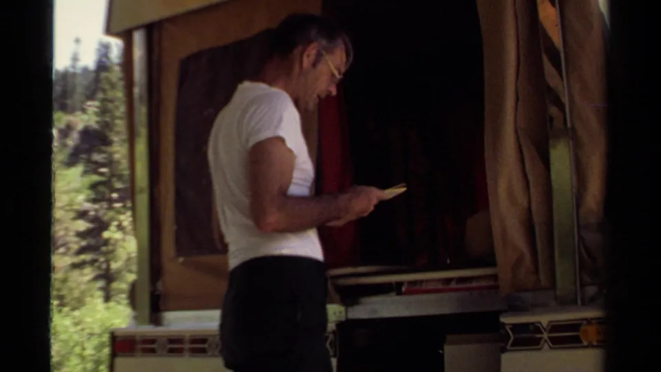 A man in a white T-shirt reads a small booklet beside an open camper trailer in Mendocino, California, 1980.