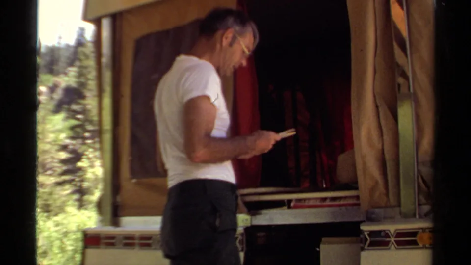 The 8mm frame shows the man reading near the open camper, with bright forest light behind the trailer.