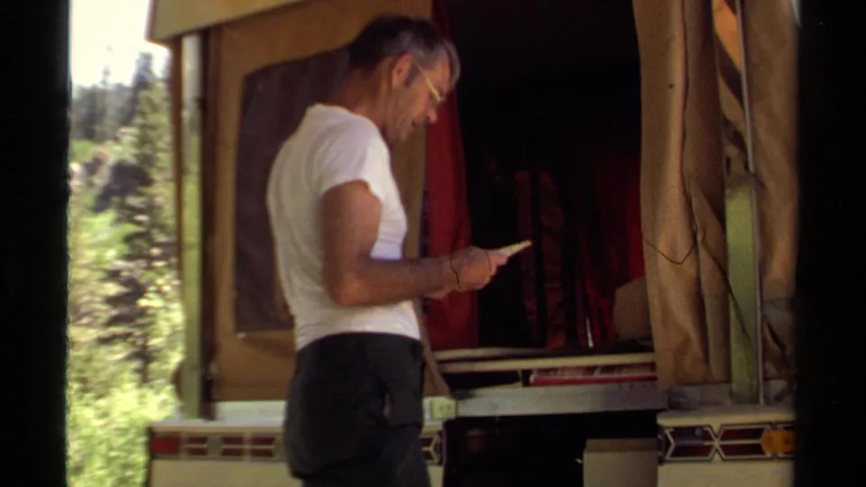 The man remains beside the open camper trailer, reading in a sunlit Mendocino forest campsite setting.