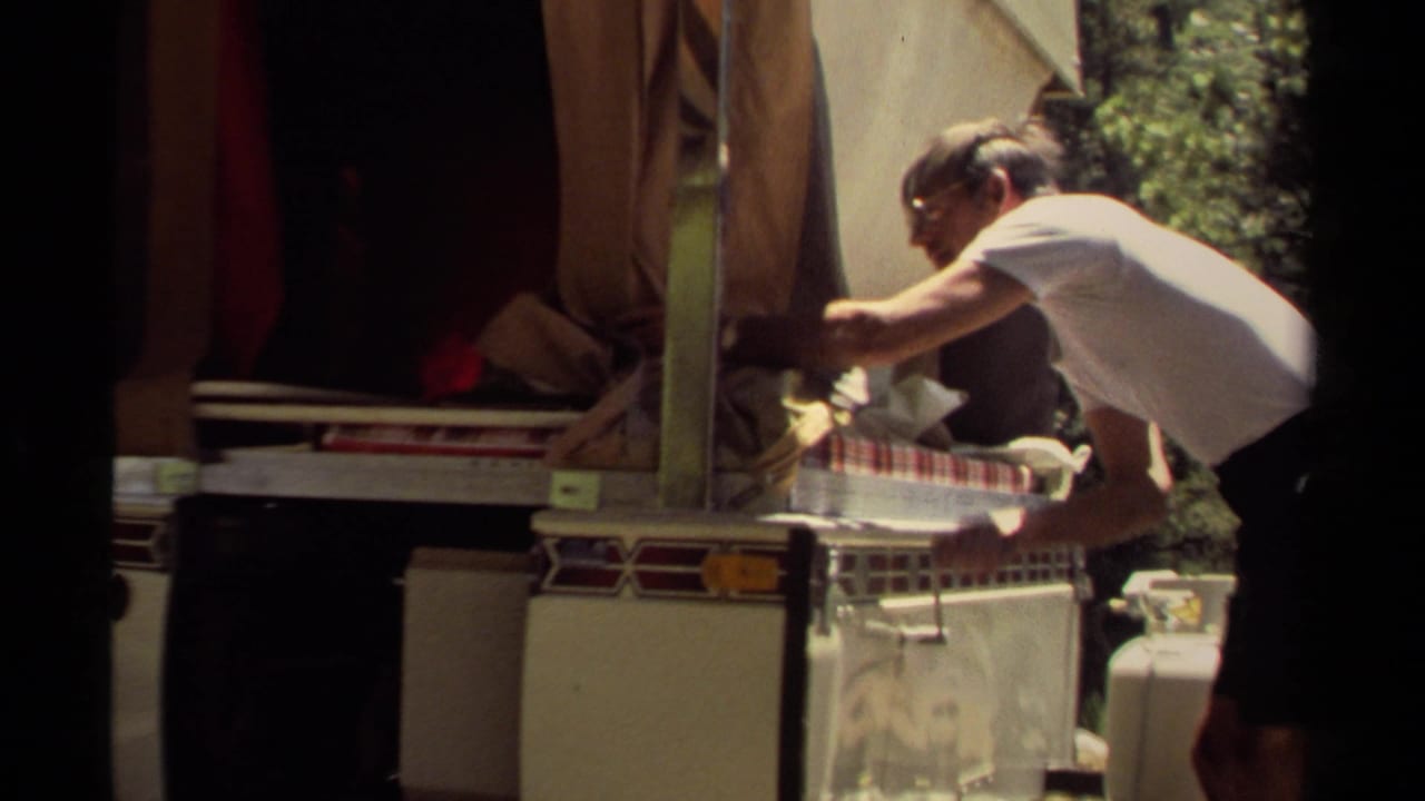 The man reaches across the open campsite trailer counter, with a checked surface, loose supplies, and a propane tank visible nearby.