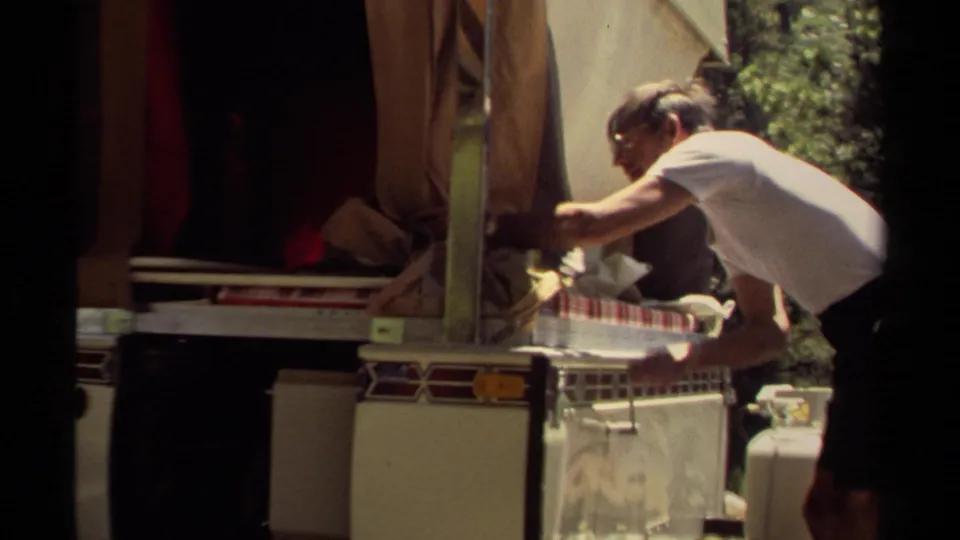 The man reaches across the open campsite trailer counter, with a checked surface, loose supplies, and a propane tank visible nearby.