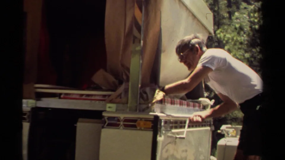 At the campsite trailer stand, the man leans in and appears to pull a hinged counter cover or support while closing the setup.