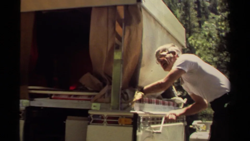 The man continues securing the small campsite food-stand counter beside the trailer, with canvas, supplies, and trees around him.