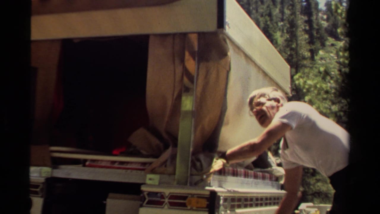 Man in a white T-shirt adjusts a camper awning beside forest trees.