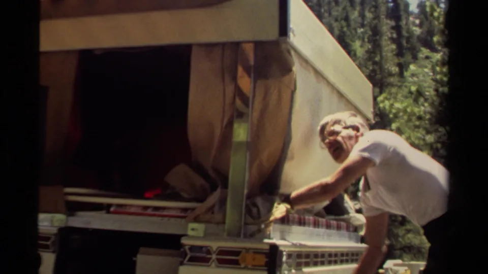 In Mendocino, California, 1980 home-movie footage, a man in glasses adjusts a canvas awning on a vintage camper in sunlit forest.