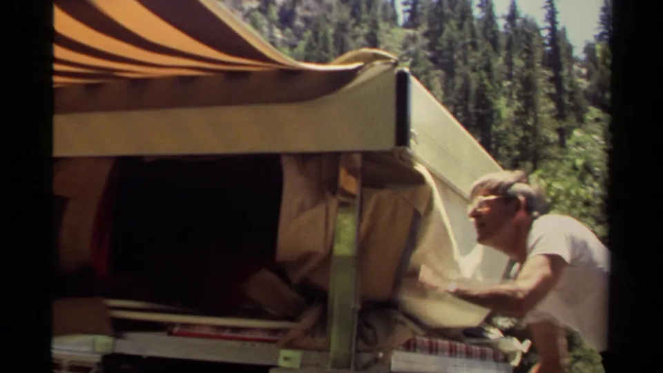 In the forest campsite scene, the man looks up while handling canvas fabric near the camper's extended awning and side panel.