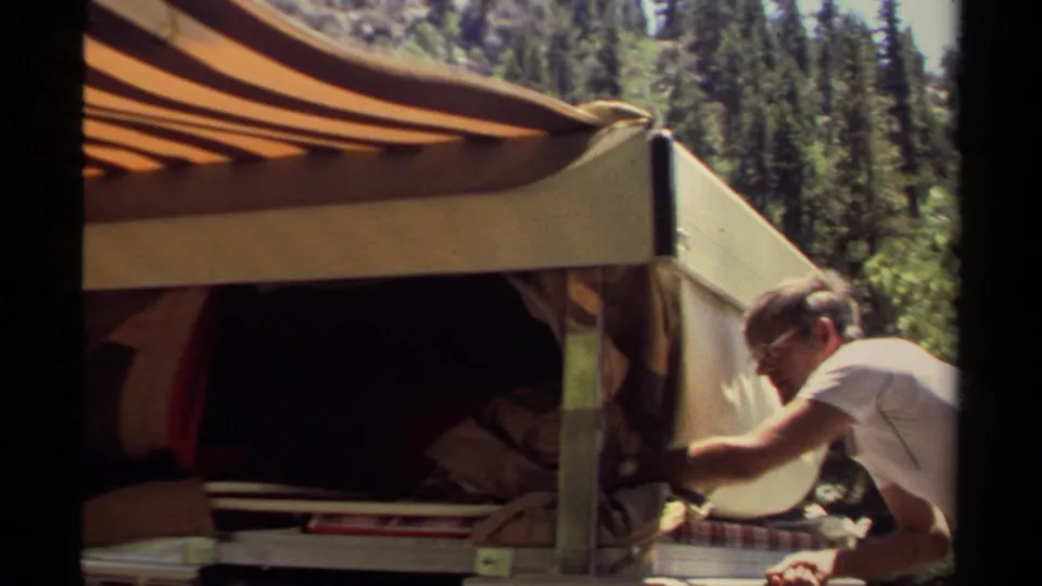 A man works at the side of a vintage camper in Mendocino, with the awning extended and pine-like trees in the background.