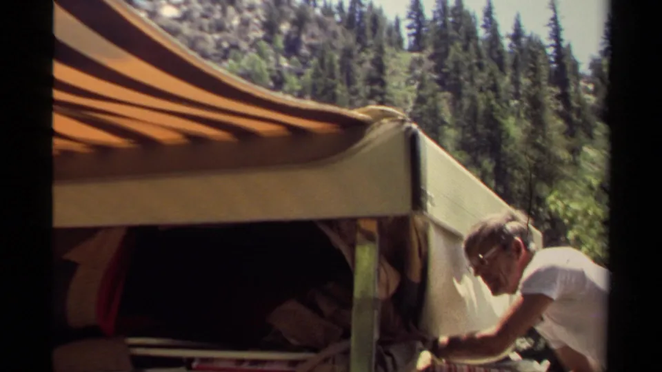 In Mendocino, California, 1980, a man works beside a vintage camper trailer with a striped awning in a sunlit forest setting.
