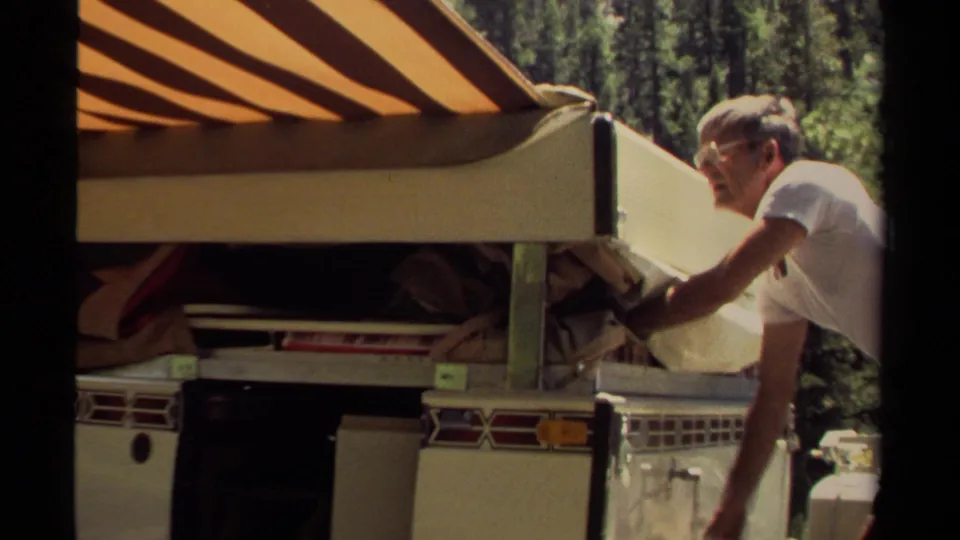 A man stands near the back of a vintage camper trailer in a forested Mendocino setting, with the striped awning and open side visible.