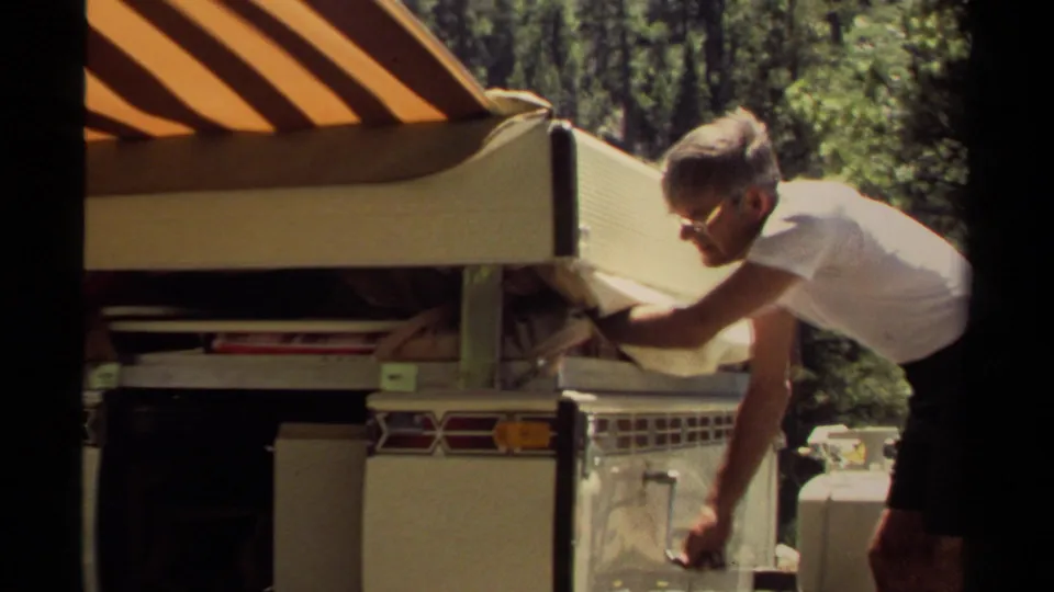 The man reaches into the open side of the camper trailer, with 1980 home-movie grain and a wooded Mendocino backdrop visible.