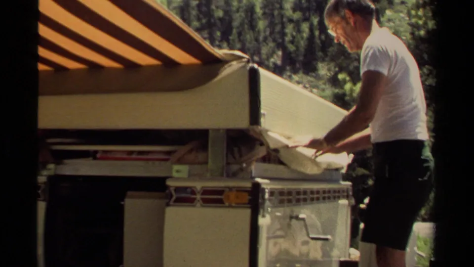 A man in a white T-shirt adjusts a light-colored camper side panel beneath a striped awning in the Mendocino forest setting.